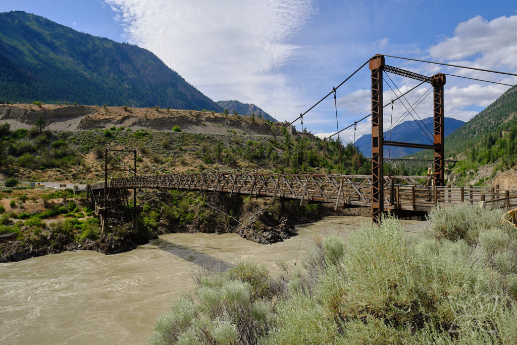 Eine alte Holzbrücke spannt sich über eine bräumlichen Fluss in einer steppenhaften Berglandschaft